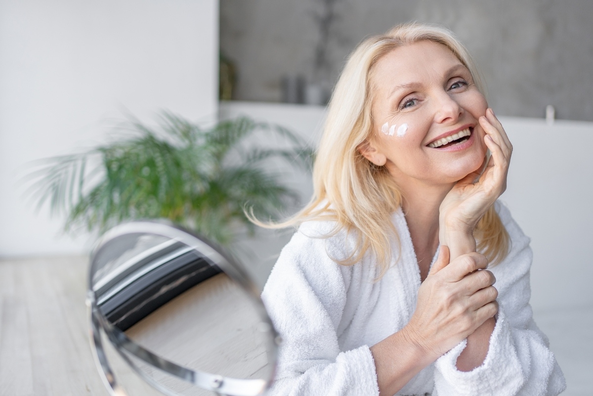 Mature woman in a white robe applying moisturizer to her face as part of XERF skin tightening aftercare following a radiofrequency treatment session
