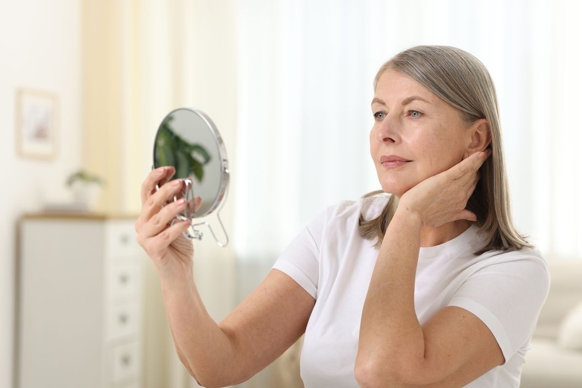 Mature woman holding a mirror and examining her jawline and neck to assess skin firmness and the right age for radiofrequency skin tightening