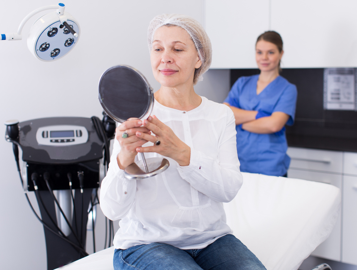 Mature woman examining her face in a mirror after a dermal filler treatment at a medical aesthetics clinic to assess how long fillers last
