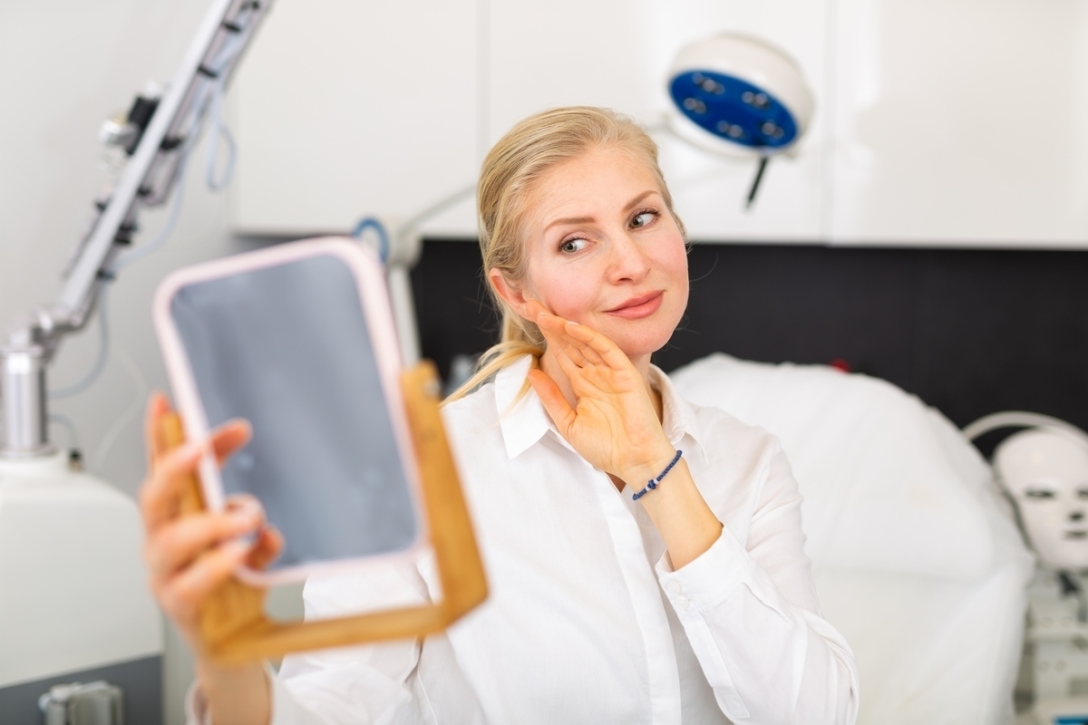 Mature woman holding a mirror and touching her cheek after a dermal filler treatment representing the post-appointment filler aftercare and results review process